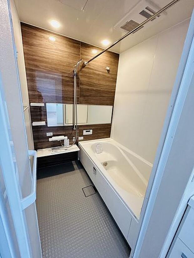 木目調のアクセントパネルが温かみを感じさせるバスルームです。広々とした浴槽でゆったりとくつろげます。天井には照明が配置されており明るい空間が広がります。壁面のミラーや収納棚も便利です。