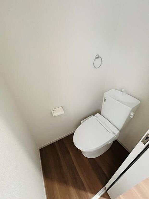 シンプルなデザインのトイレで白を基調とした清潔感のある空間です。木目調の床が落ち着いた印象を与えます。壁面にはペーパーホルダーやタオルリングが配置されています。ドアの開閉もスムーズです。