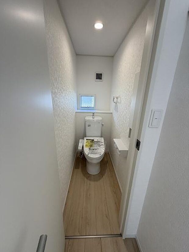 ナチュラルな木目調の床が印象的なトイレです。天井のダウンライトで明るく清潔感のある空間になっています。小窓が配置されており採光や換気にも配慮されています。壁の質感もアクセントになっています。