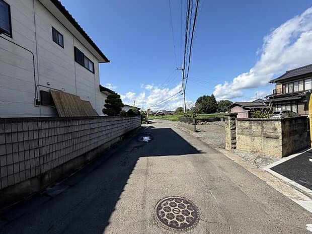 前面道路は舗装されており車両の出入りがしやすい環境です。周囲には住宅が並び整然とした街並みが特徴です。空が広く感じられ開放感があります。道路沿いにはブロック塀や石垣が整然と配置されています。
