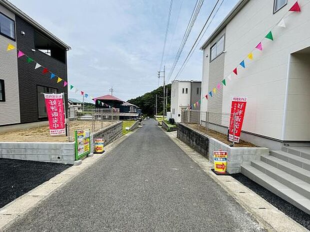 現地写真では前面道路が広がり両側に新しい住宅が並んでいる様子が分かります。電柱やカラフルな旗が設置されていて周囲は落ち着いた雰囲気です。道路の状態も良好で日当たりも期待できる環境となっています。