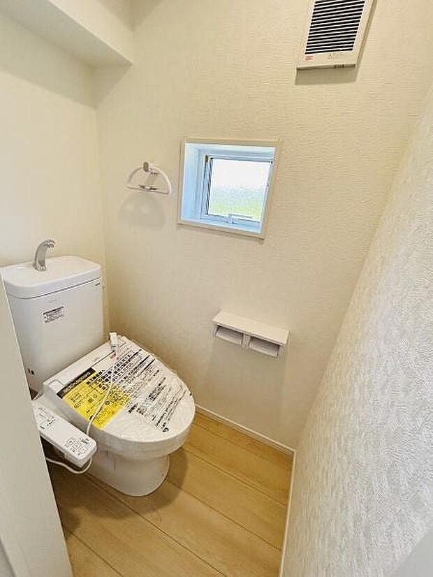 窓から光が差し込むトイレです。壁付けタイプのトイレットペーパーホルダーが設置されています。温水洗浄便座が付いており快適に使用できます。コンパクトながらも換気設備が整っています。
