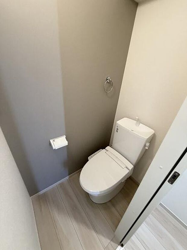 シンプルなデザインのトイレ空間です。落ち着いた色合いの壁面が特徴的です。ペーパーホルダーとタオルリングも備わっています。床は明るめの色で清潔感があります。扉の開閉もスムーズに行えます。