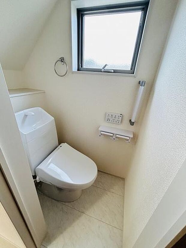 窓が設置されており自然光が入る明るいトイレです。壁付けのペーパーホルダーや操作パネルが備わっています。スペースを有効に活用したシンプルな内装です。床や壁も手入れがしやすい仕様になっています。