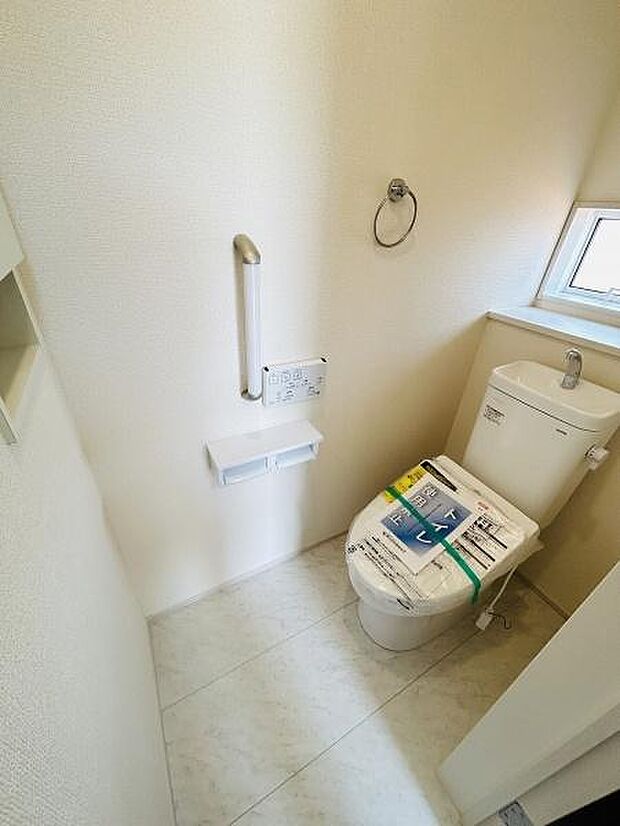 窓から自然光が入る明るいトイレです。手すりが設置されており使いやすさにも配慮されています。シンプルな内装で清潔感のある空間です。ペーパーホルダーやタオルリングも備わっています。