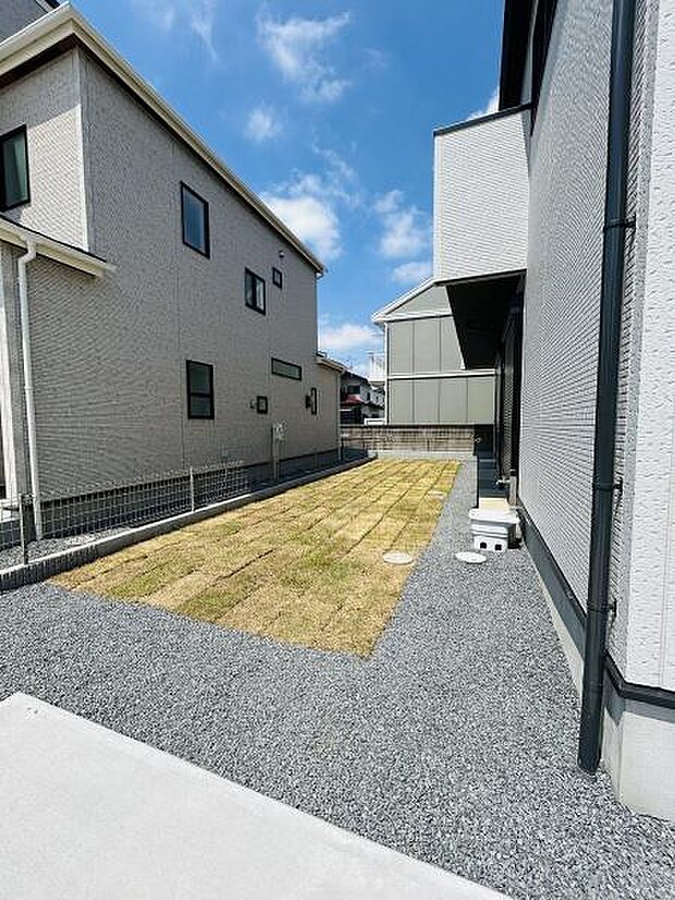 手入れのしやすい芝生スペースが広がる庭です。両側の建物に囲まれておりプライベートな空間が確保されています。明るい空の下シンプルな外構が印象的です。使い勝手のよい動線も確保されています。