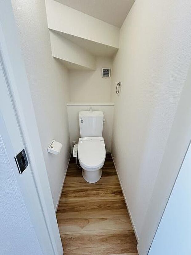 ホワイトを基調としたシンプルなトイレ空間です。床材が木目調で温かみを感じられます。壁面にペーパーホルダーやタオルリングが設置されています。居心地の良いスペースが広がっています。清掃もしやすい造りです。