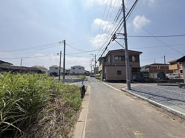 周囲には住宅が並び落ち着いた住宅街の雰囲気が感じられます。前面道路は舗装されていて通行しやすい環境です。電柱や電線がしっかり整備されておりインフラ面も安心です。晴れた空が広がり道沿いに緑が見られます。