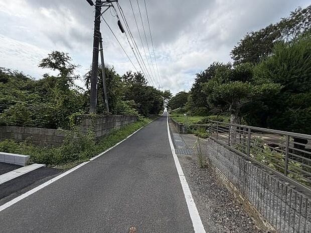 前面道路は見通しが良く周囲には緑豊かな自然が広がっています。道路幅もしっかり確保されているため通行もしやすい環境です。落ち着いた雰囲気が感じられる現地写真です。道路脇のフェンスや植栽も整っています。