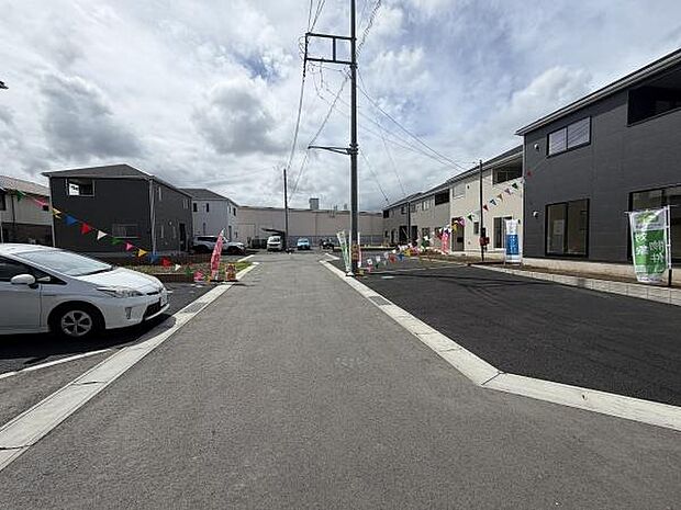 前面道路を含む現地写真です。舗装された道路が広がり車の出入りもしやすい設計が特徴です。両側には新しい住宅が並んでおり周辺環境も整っています。旗や案内が設置されており明るい印象です。