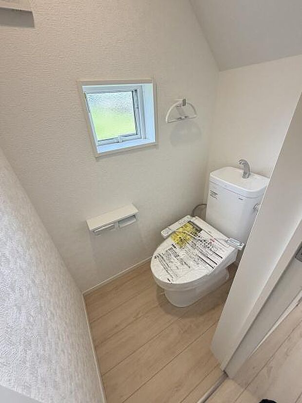 窓付きで換気しやすいトイレ空間です。シンプルな内装で清潔感があります。手洗い付きのタンクが設置されています。床は明るい色合いで圧迫感を抑えています。ペーパーホルダーも壁付けで使いやすい設計です。