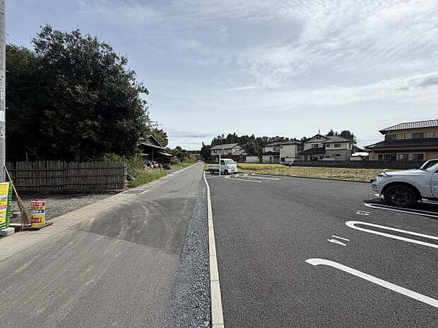 舗装された前面道路が広がり落ち着いた雰囲気の現地写真です。周囲には住宅や緑が点在し住環境を感じられます。駐車スペースも見られるため利便性が伺えます。ゆとりある道路幅が印象的です。