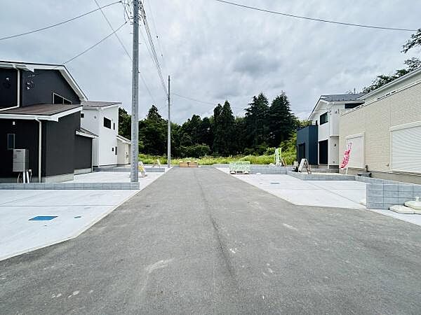 広々とした前面道路が印象的な現地写真です。両側にゆとりのあるスペースが確保されています。周辺に緑も感じられる静かな環境です。建物周辺の舗装も丁寧に整っています。