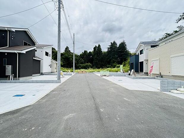 広々とした前面道路が印象的な現地写真です。両側にゆとりのあるスペースが確保されています。周辺に緑も感じられる静かな環境です。建物周辺の舗装も丁寧に整っています。