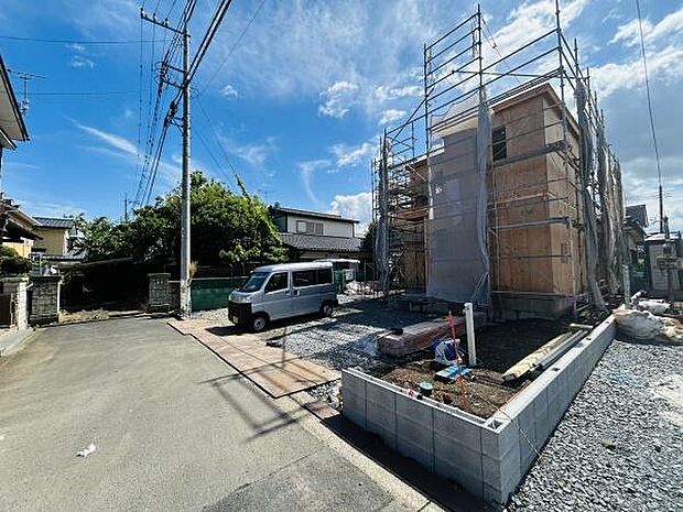 建築中の物件が立ち並ぶ現場です。敷地周辺には広々としたスペースが確保されています。前面道路がゆったりとしており車の出し入れも便利です。晴天のもと開放的な印象を受けます。