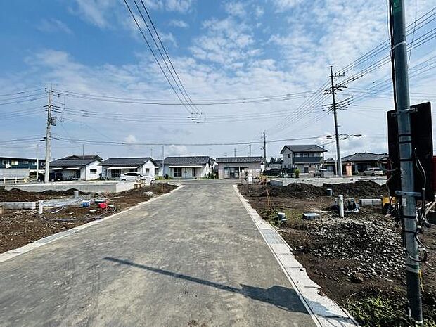 前面道路が広く見通しの良い現地写真です。周囲には新しい住宅や建物が整然と並んでいます。空が広く開放感があります。道路は舗装されており整備が進んでいます。