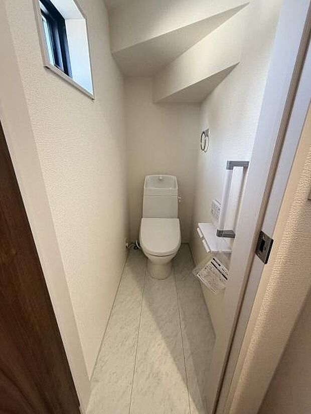 シンプルで清潔感のあるトイレ空間です。コンパクトな設計で使いやすさに配慮されています。窓があり換気しやすい設計です。手すりや収納スペースも設置されています。壁や床の色調も落ち着いています。
