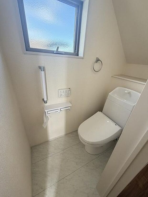 窓付きで明るさを感じられるトイレ空間です。手すりやペーパーホルダーが設置されており使いやすさも追求されています。シンプルな壁面で掃除もスムーズです。収納棚が設けられており物の整理も便利です。