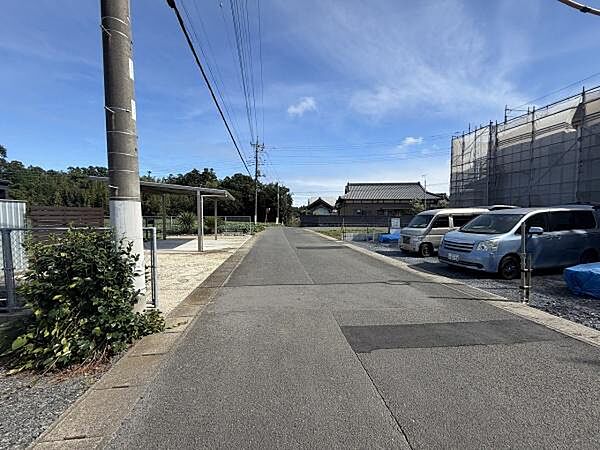 前面道路は舗装されており周囲に駐車スペースが整っています。電柱やフェンスも整然と配置されています。空が広く感じられ周辺には住宅や建物が配置されています。見通しの良い道路環境となっています。