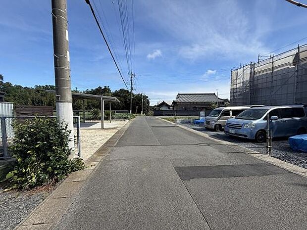 前面道路は舗装されており周囲に駐車スペースが整っています。電柱やフェンスも整然と配置されています。空が広く感じられ周辺には住宅や建物が配置されています。見通しの良い道路環境となっています。