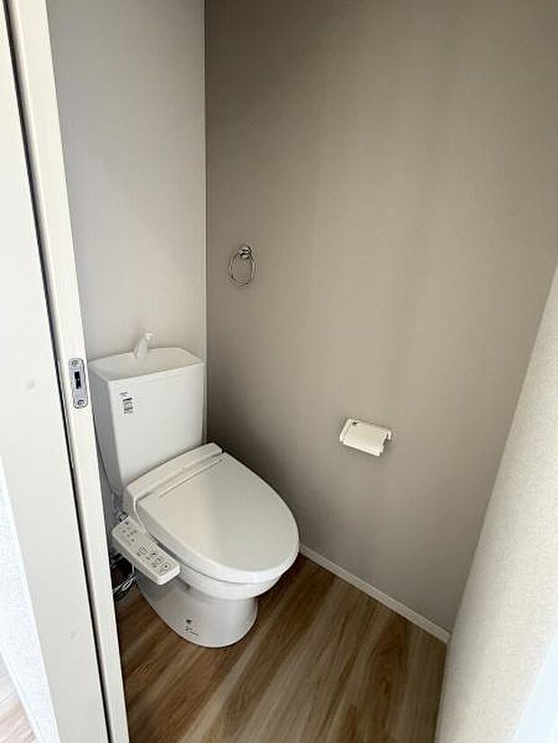 シンプルなデザインのトイレで清掃がしやすい仕様です。落ち着いた色味の壁と床が空間に調和しています。コンパクトながらも必要な設備が整っています。ペーパーホルダーやタオルリングも設置されています。