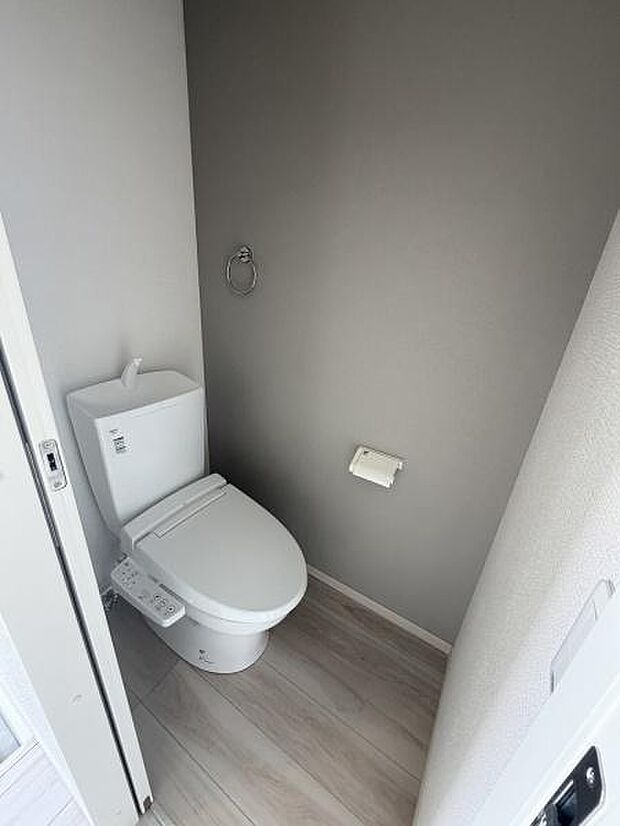 グレーのアクセントクロスが印象的なトイレです。シンプルなデザインで落ち着いた雰囲気が漂います。床材とのコントラストが空間を引き締めています。ペーパーホルダーやタオルリングも備わっています。