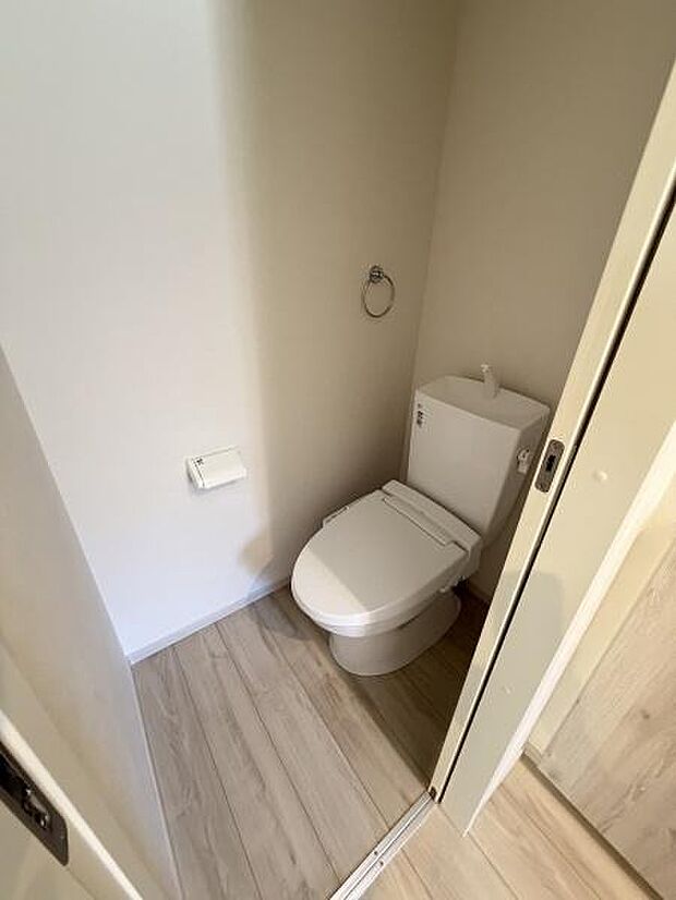 ナチュラルな木目調の床が落ち着いた雰囲気を演出しています。シンプルなホワイトのトイレが空間をすっきりと見せています。タオルリングやペーパーホルダーも設置されており使い勝手に配慮されています。