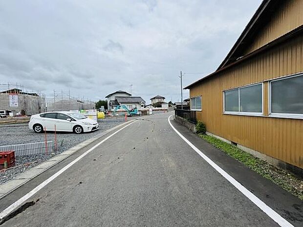 現地前面道路はゆるやかなカーブが特徴的です。周辺は落ち着いた住宅が並んでいます。道幅もしっかり確保されており駐車スペースや歩行者にも配慮されています。現地の環境が分かりやすい写真です。