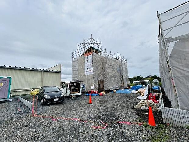 現在工事中の建物で全体がしっかりと養生されている様子が見られます。広い敷地には車や資材が配置され現場の整理が感じられます。安全対策としてコーンも設置されています。駐車スペースも確保されています。