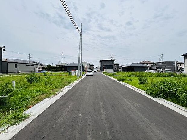 前面道路は広く車の出入りがスムーズに行えます。周囲には住宅が並び整然とした街並みが広がっています。道路の舗装も新しく快適な通行が可能です。周囲の緑も感じられる現地写真です。