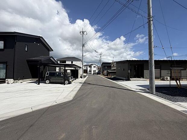 前面道路が広く見通しの良い現地写真です。舗装された道路が清潔感を演出しています。周辺には新しい建物が建ち並び整った印象を受けます。電柱や配線も整然としており安心感があります。