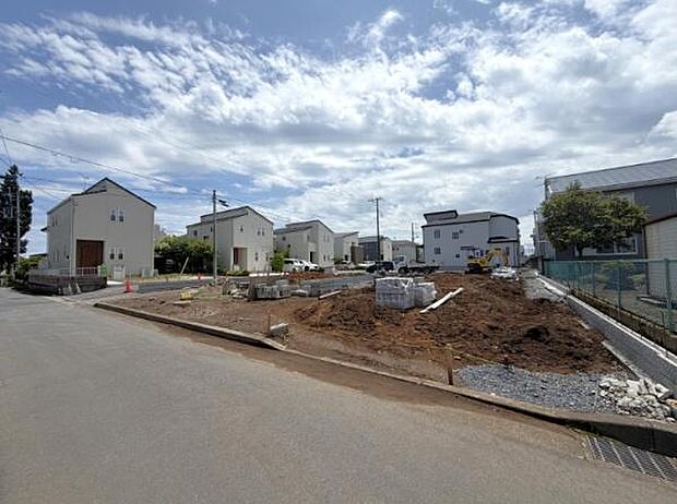 前面道路が広く車の出し入れがしやすい現地写真です。周囲には住宅が並び整った街並みが見受けられます。開放感のある現地風景が印象的です。建築が進む様子も確認できます。空の広がりが感じられます。