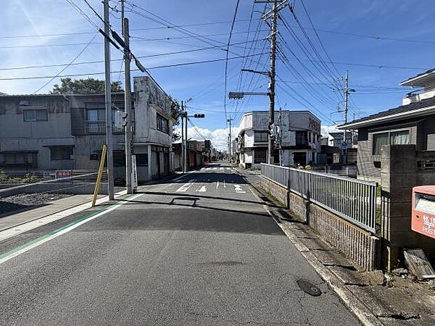 前面道路は幅員が広く見通しのよい直線道路が続いています。舗装の状態も良好で交通標識も設置されています。周辺は住宅や商業施設が並び整然とした印象です。電柱やフェンスなども整備が行き届いています。