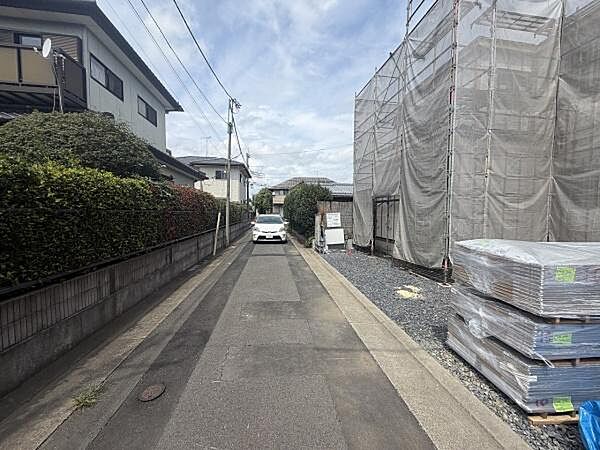 前面道路は舗装されており周囲には住宅が立ち並んでいます。落ち着いた雰囲気の住宅街の一角です。現地敷地には資材が置かれており工事中の様子が見受けられます。電柱や配線も整然と配置されています。