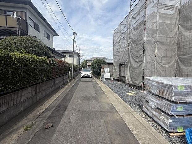 前面道路は舗装されており周囲には住宅が立ち並んでいます。落ち着いた雰囲気の住宅街の一角です。現地敷地には資材が置かれており工事中の様子が見受けられます。電柱や配線も整然と配置されています。