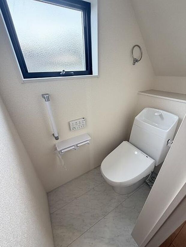 窓付きで換気がしやすいトイレです。清潔感のある白い内装が特徴です。手すりや温水洗浄便座も備えています。ペーパーホルダーや小物掛けも設置されています。棚付きで収納も便利です。