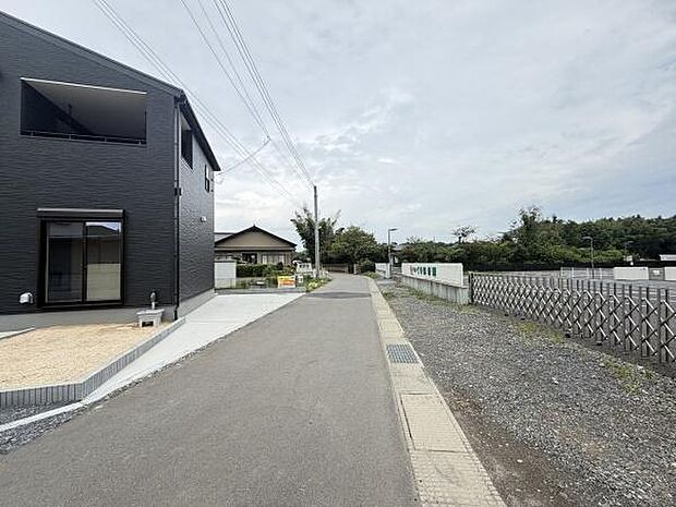 前面道路がすっきりと整備されており車の出入りがしやすい環境です。周辺には緑も多く開放感のある現地写真です。歩道も確保されていて日々の移動に安心感があります。ゆったりとした道路幅が印象的です。