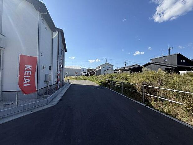 前面道路は舗装されておりきれいな状態です。道路幅にゆとりがあり車の通行も安心です。周囲には新しい建物が並んでおり落ち着いた雰囲気です。道路脇にはフェンスも設置されています。