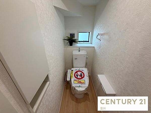 白を基調とした清潔感のあるトイレ空間です。温水洗浄便座や手洗いカウンターを備えているほか、小窓からの採光で明るく、サイドには棚付きの収納スペースも設置されています。