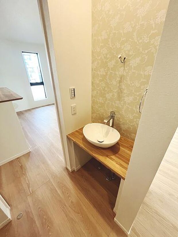 シンプルなデザインの手洗いスペースがトイレ横に設けられています。木目調のカウンターが温もりを感じさせます。明るい壁紙が空間を柔らかく演出しています。毎日の使用にも配慮された設計です。