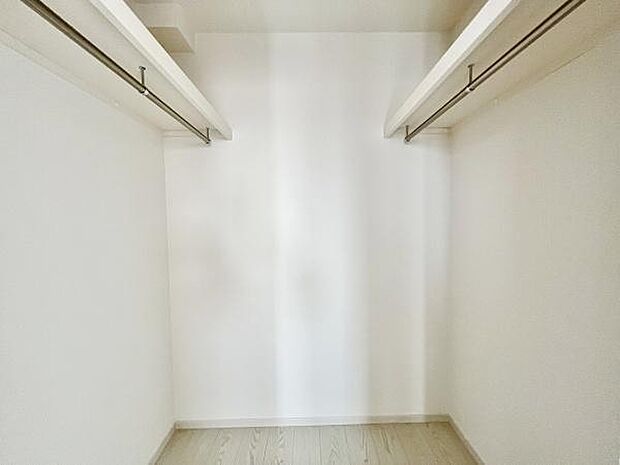 シンプルなデザインの収納スペースです。清潔感のある内装が特徴です。ハンガーパイプが両側に備え付けられています。ホワイトを基調とした空間です。床にはナチュラルな色合いのフローリングが採用されています。