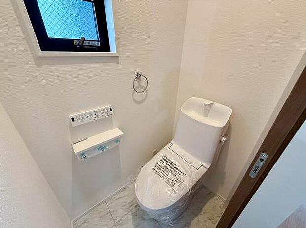 シンプルで清潔感のあるトイレ空間です。手洗い一体型の便器が備わっています。壁紙や床も明るい色でまとめられ開放感があります。収納やペーパーホルダーなども設置されています。