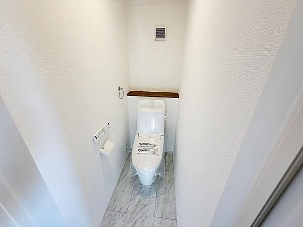 白を基調とした清潔感のある空間です。壁付けのペーパーホルダーが設置されています。上部棚には小物を置けるスペースがあります。床は石目調の仕上げがされています。通気口があり換気もしやすくなっています。