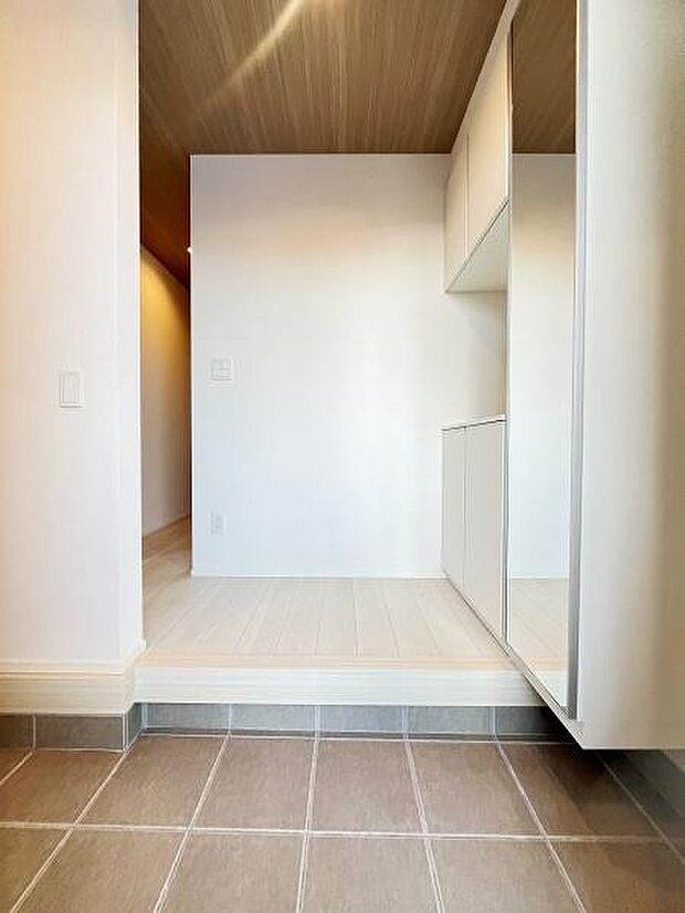 シンプルなデザインが目を引く玄関です。落ち着いた色合いの床タイルが清潔感を演出しています。収納スペースも備わっています。天井の木目調がアクセントになっています。