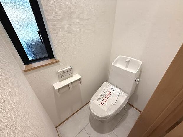 シンプルなデザインのトイレ空間です。窓付きで自然光が差し込みます。操作しやすいウォシュレット付きリモコンが備わっています。床はお手入れしやすい仕様です。清潔感を感じられる内装です。