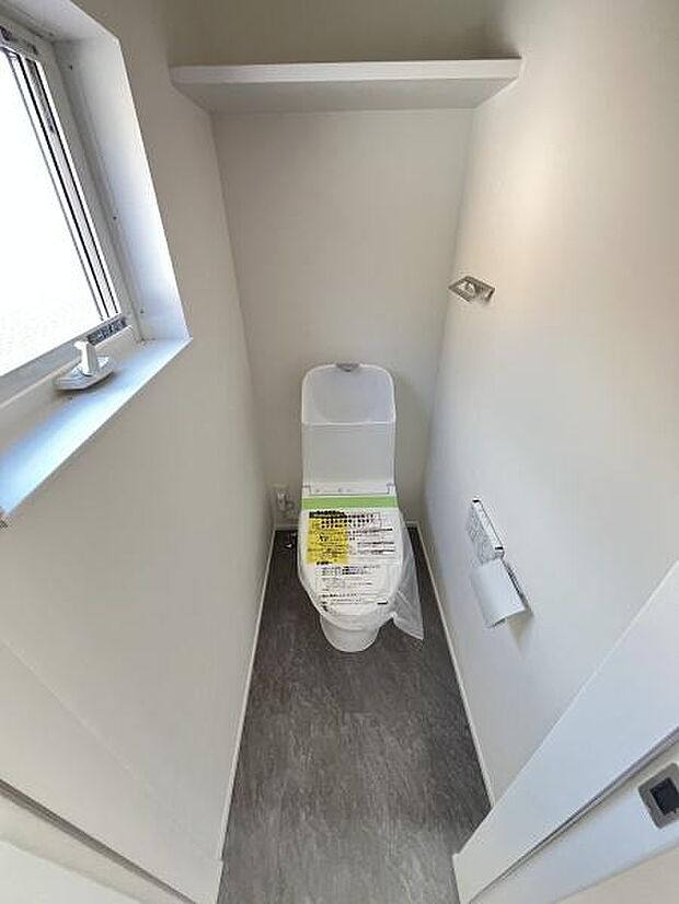 清潔感のあるシンプルな内装のトイレです。窓付きで換気がしやすく明るい空間が印象的です。棚も設置されており小物の収納に便利です。床はお手入れしやすい仕様となっています。