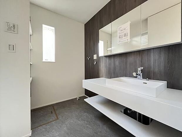 洗面台がシンプルなデザインで清潔感が際立ちます。鏡裏に収納スペースが設けられており使い勝手も考慮されています。窓から自然光が入り空間が明るく見えます。壁面のダークカラーが全体を引き締めています。