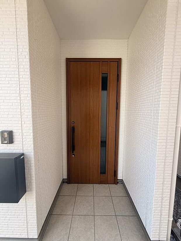 玄関の木目調ドアがシンプルで落ち着いた印象を与えます。白い外壁とタイル床がすっきりとした雰囲気を演出しています。ポストやインターホンも設置されており使い勝手の良さが感じられます。