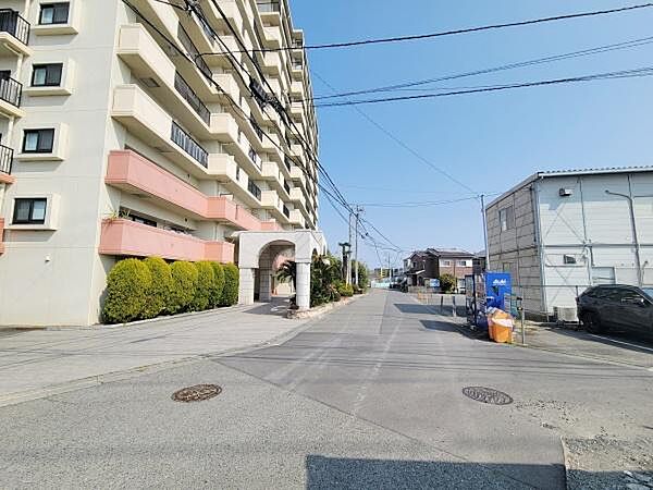 明るい空の下で清潔感のあるマンション外観が印象的です。エントランス前には緑の植栽が整えられており通りも開放的です。周囲は道幅が広く車の出入りもしやすい環境です。