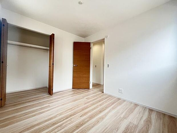 洋室はナチュラルな色合いのフローリングが特徴的です。大型のクローゼットが設置されており収納スペースも確保されています。室内は白を基調とした壁面で明るい印象です。ドアや収納扉は木目調で統一されています。